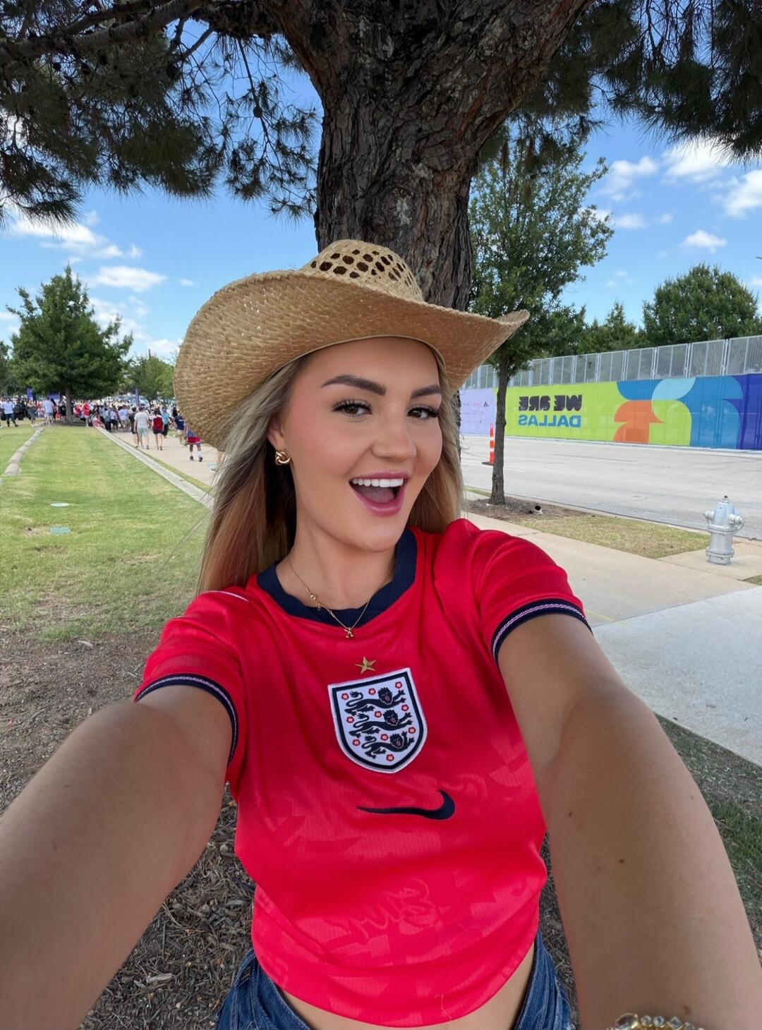 A young woman in a red England football jersey and a straw cowboy hat takes a selfie outdoors. She is smiling broadly. In the background, there's a tree, green grass, people walking, and a wall with 'WE SEE DALLAS' written on it (mirrored).