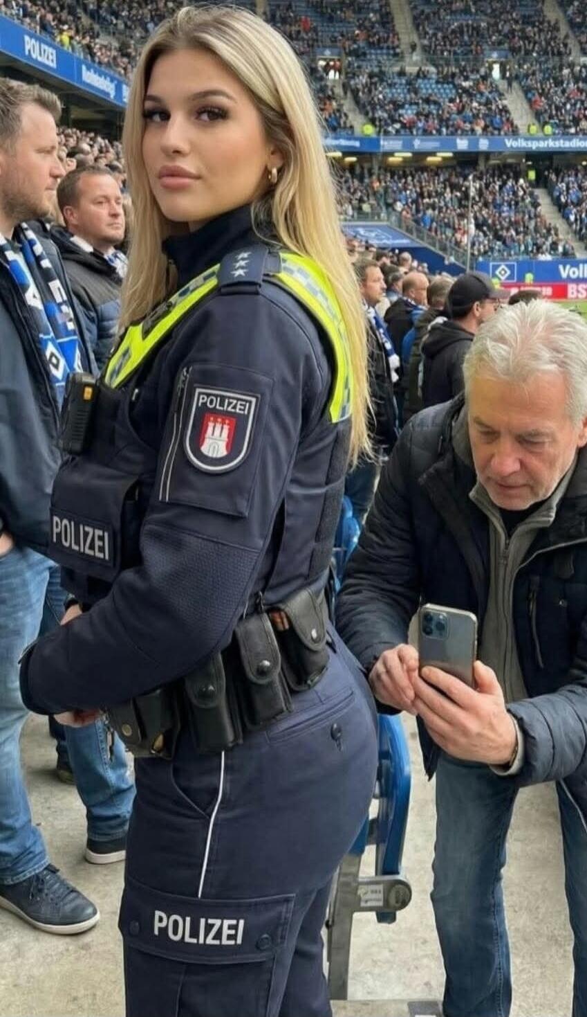 POLIZEI, Volksparkstadion. A female police officer in uniform is standing at a stadium, looking towards the camera. An older man next to her is holding a smartphone, seemingly taking a picture of her from behind.