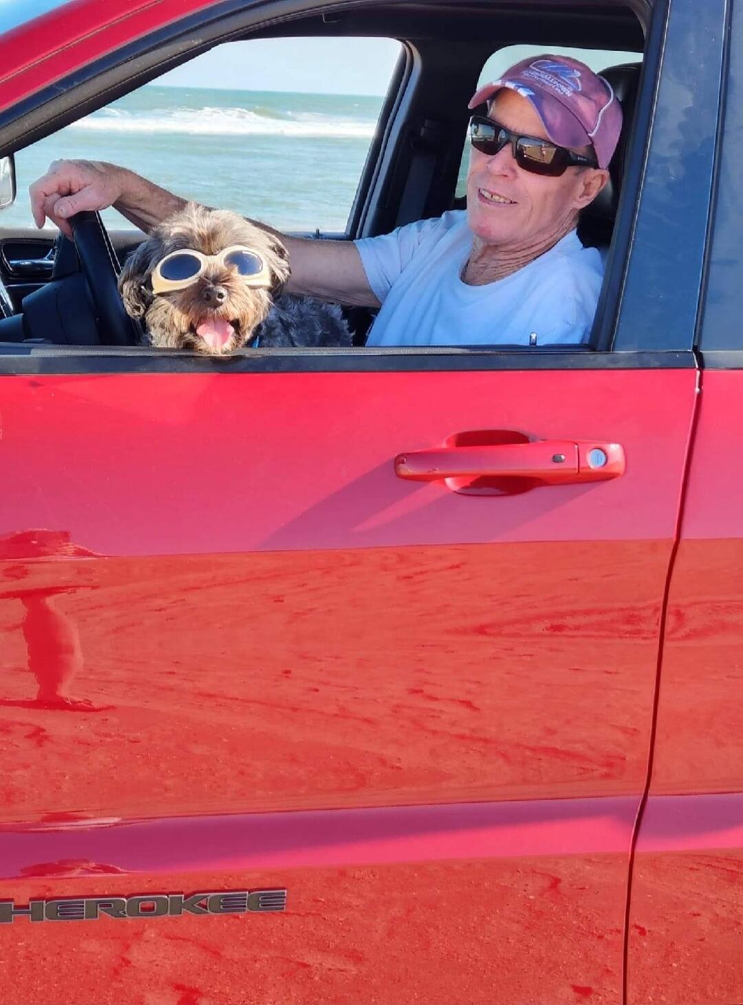 The man and the dog are in a red car. The dog is wearing sunglasses.