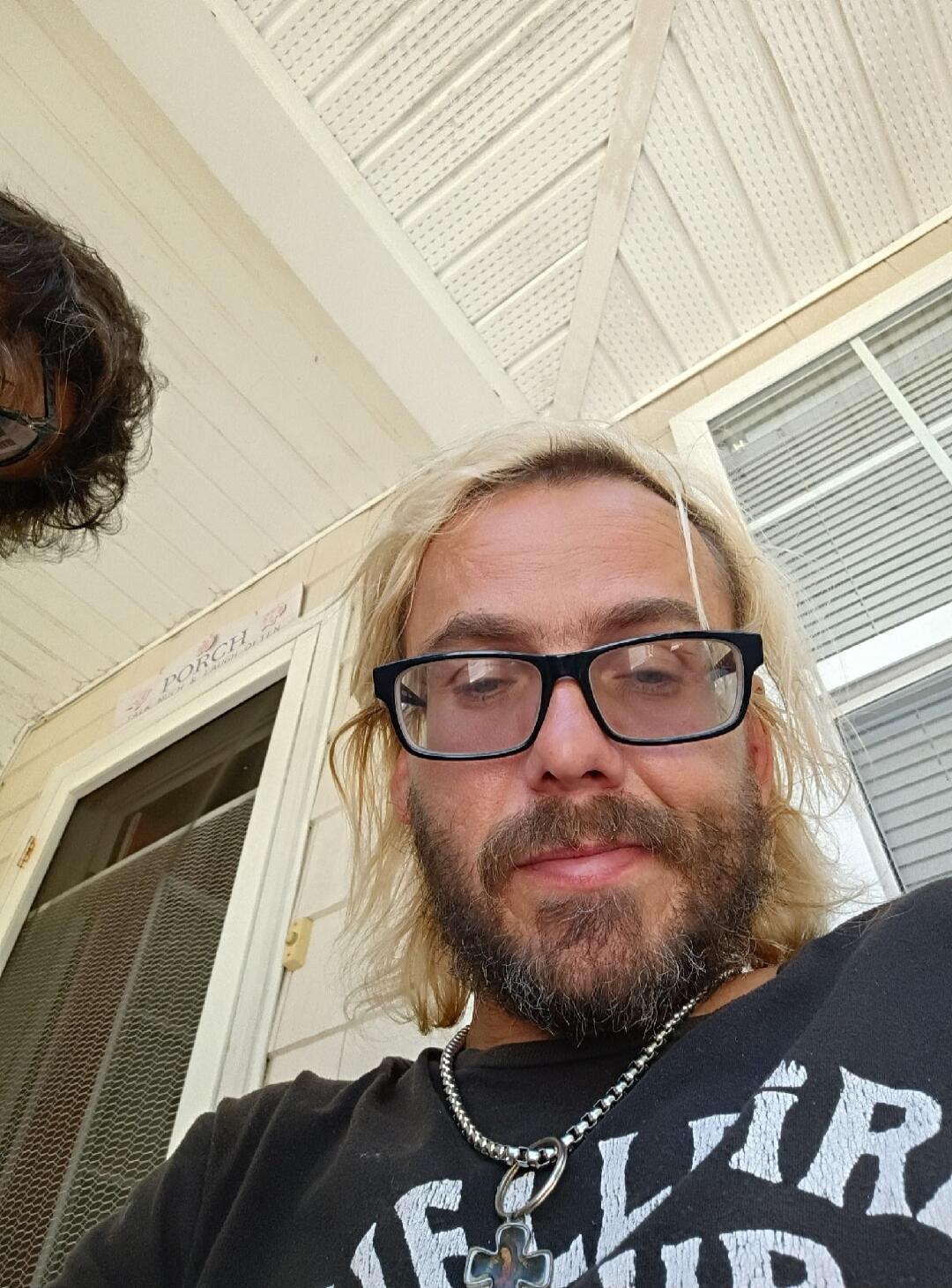 PORCH TALK MUCH & LAUGH OFTEN. A man with blonde hair, a beard, and glasses takes a selfie. Another person's head is partially visible on the left. They appear to be on a porch.