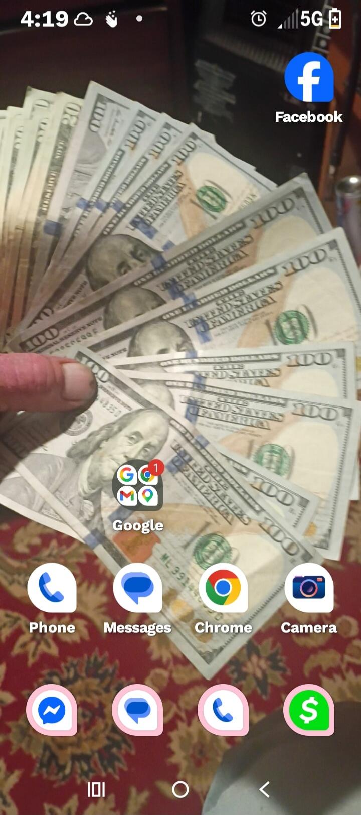 A hand holds a fan of one hundred dollar bills, partially covering a smartphone screen displaying app icons such as Facebook, Google, Phone, Messages, Chrome, and Camera. The time 4:19 and 5G network indicator are visible at the top of the screen. The bills show 'UNITED STATES OF AMERICA' and 'ONE HUNDRED DOLLARS'.