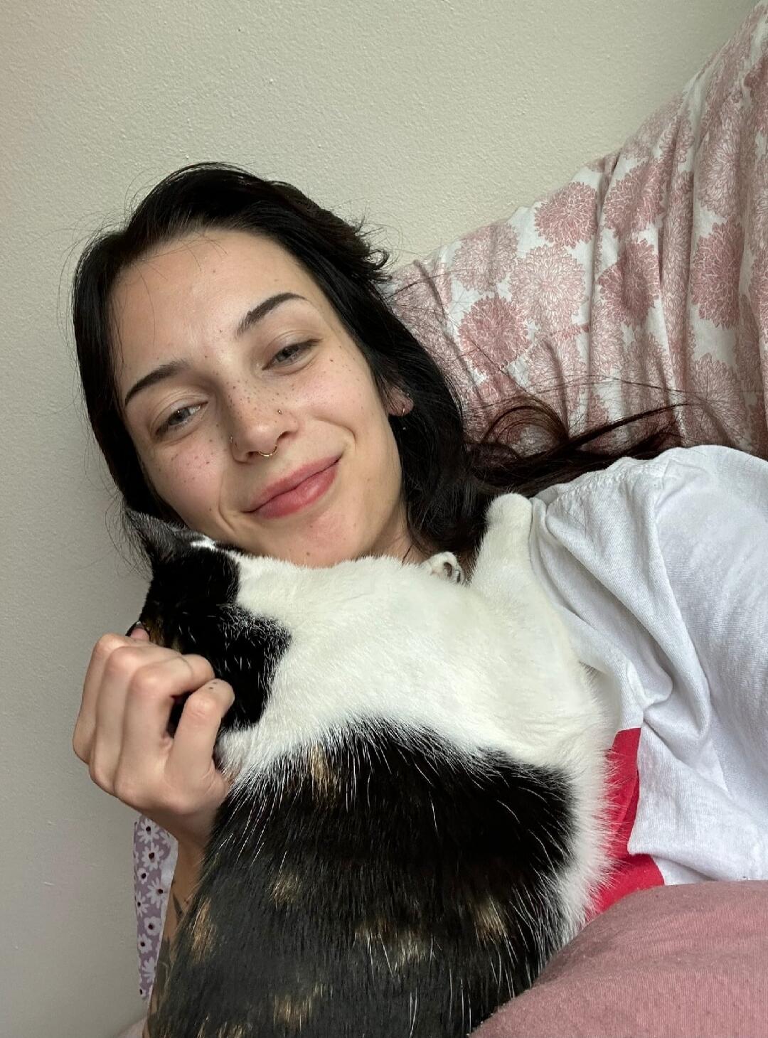 A young woman with dark hair and freckles is smiling while cuddling a calico cat. She appears to be lying down comfortably.