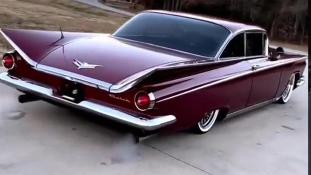 A classic burgundy car with prominent tail fins and white wall tires, possibly a Buick from the late 1950s, is shown from the rear. Some smoke is visible coming from the exhaust pipe.