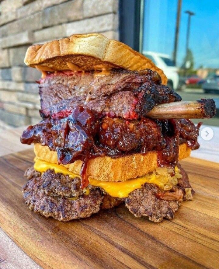 This is a massive, multi-layered sandwich, likely a burger or a barbecue creation. It features thick slices of toasted bread, generously filled with layers of meat. The visible components include what appears to be slow-cooked brisket or ribs with a bone sticking out, covered in a rich, dark barbecue sauce. Beneath this, there are slices of melted 