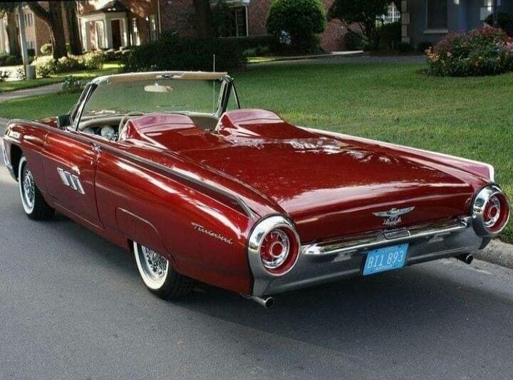 A classic red Ford Thunderbird convertible car is parked on a residential street. The car has white wall tires and chrome accents. The license plate reads BII 893. The background shows green lawns and houses.