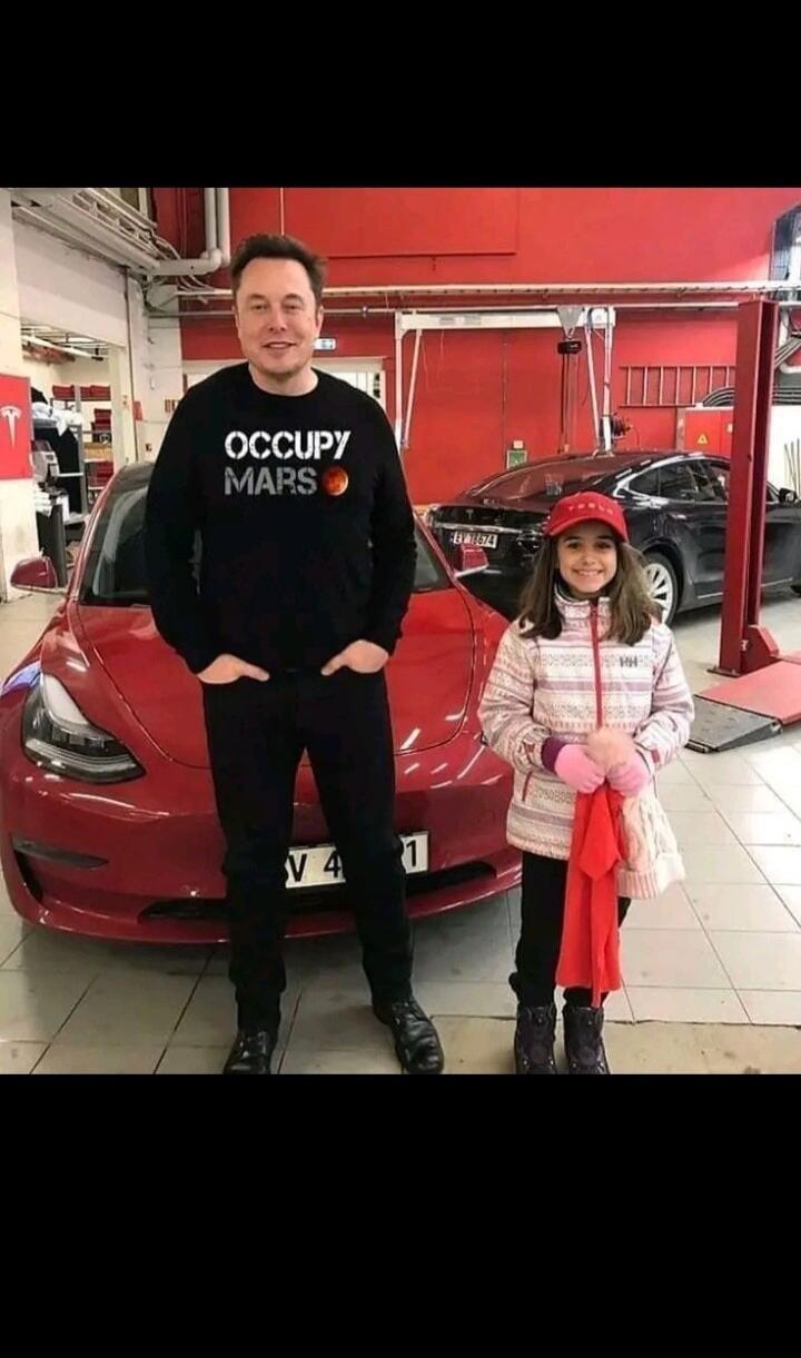 Elon Musk wearing an 'OCCUPY MARS' shirt stands next to a red Tesla car with a young girl. Another black Tesla is visible in the background of what appears to be a service station or garage.