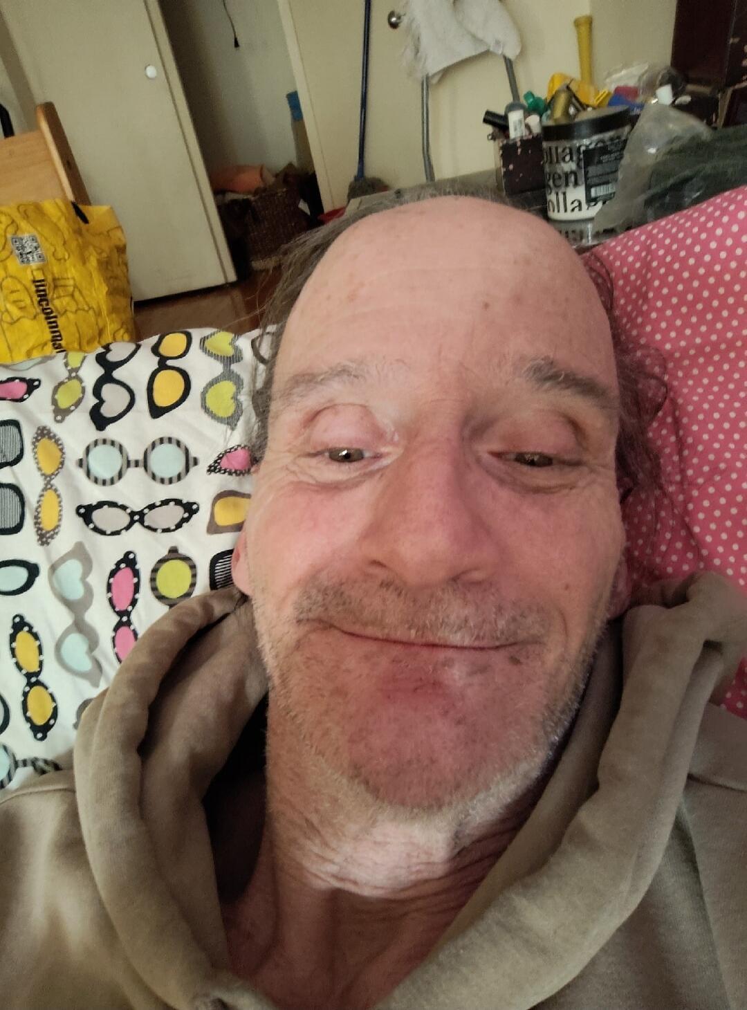 A man taking a selfie while lying down. Text on a yellow bag reads 'Lincolnmal'. Text on a container in the background reads 'en olla'.