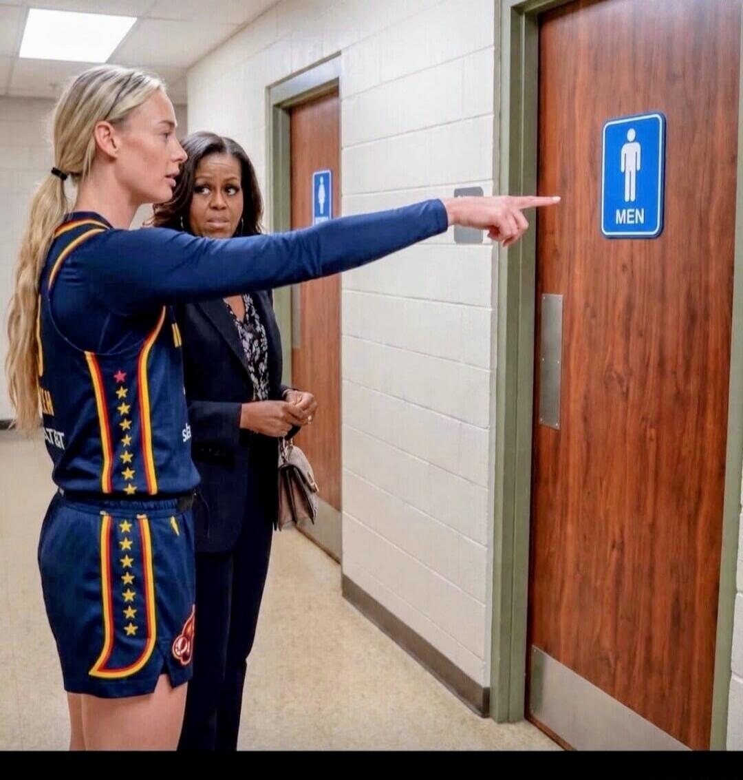 A blonde woman in a basketball uniform points to a men's restroom sign while Michelle Obama looks on with a surprised expression. The sign clearly indicates 'MEN'.