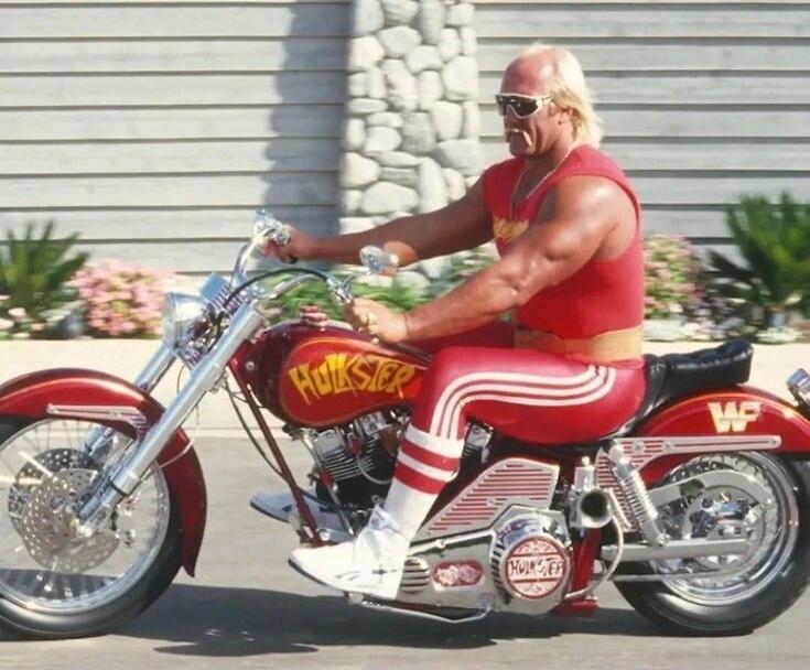 Hulk Hogan riding a custom red motorcycle. The motorcycle has 'HULKSTER' written on the tank and engine cover, and 'WF' on the rear fender. He is wearing his signature red and white wrestling attire and sunglasses.