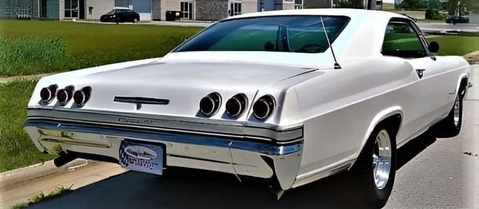 A white classic car, possibly a Chevrolet Impala, is parked on an asphalt surface with a grassy area and buildings in the background.