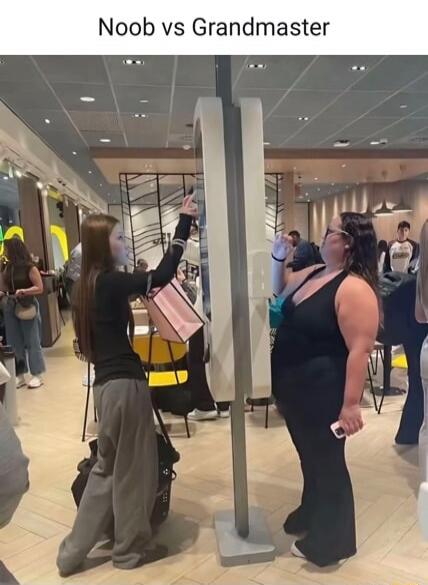 Noob vs Grandmaster. Two women are taking photos or selfies in a public space. One woman is awkwardly holding her phone high, seemingly struggling to find the right angle, while the other woman stands more casually, holding her phone lower and appearing more confident in her photo-taking technique.