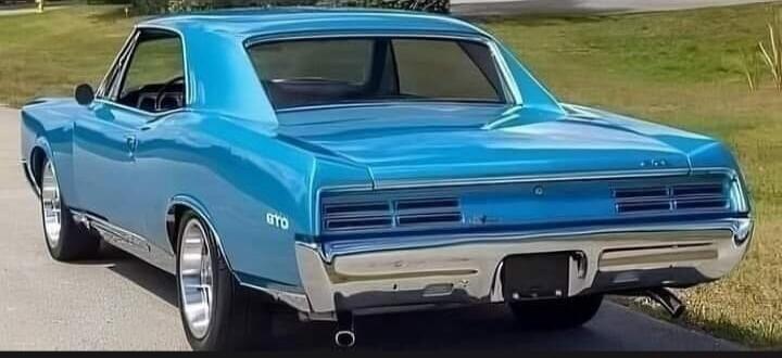 A classic blue Pontiac GTO muscle car, seen from the rear-side, parked on an asphalt road next to a grassy area. The car features chrome bumpers and custom wheels.