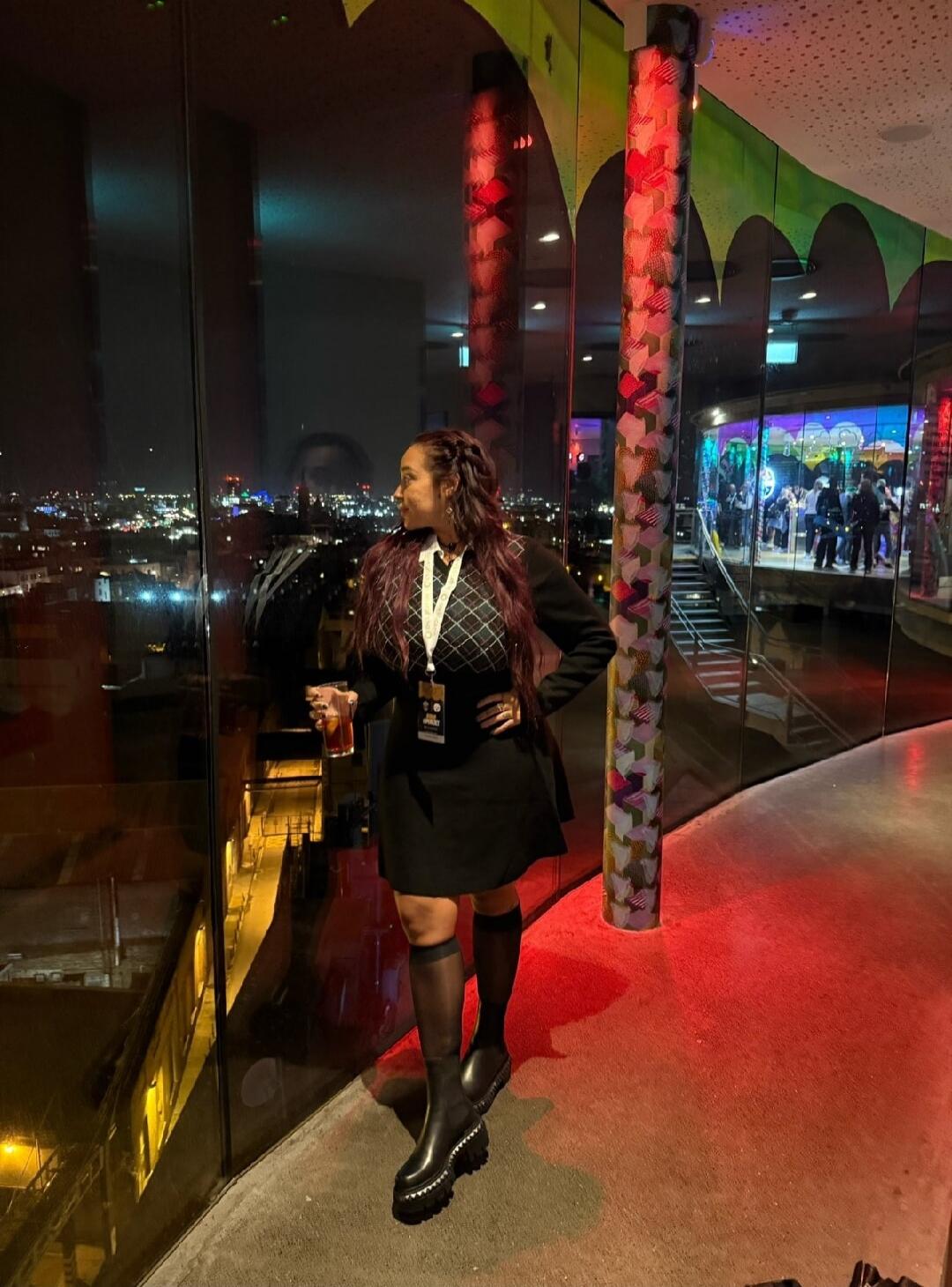 A woman in a black dress with a white collar and a patterned top is standing by a large window at night, holding a drink. She is looking out at the illuminated city skyline. She is wearing a lanyard with a badge. The interior features red lighting on the floor and decorative pillars. A reflection of another person is visible in the window.