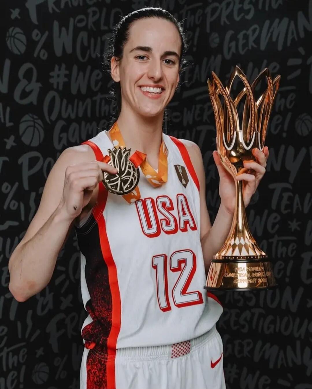 A female basketball player wearing a USA jersey with number 12, holding a gold medal and a golden trophy. The trophy is from the FIBA Women's Basketball World Cup Germany 2018. The background includes text like 'WORLD CUP', 'WE OWN THE GAME', 'GERMANY', and 'THE PRESTIGIOUS'.