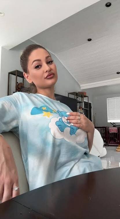 A woman with her hair pulled back is sitting at a table, wearing a light blue tie-dye long-sleeve shirt with a cartoon character on it. She is looking directly at the camera.