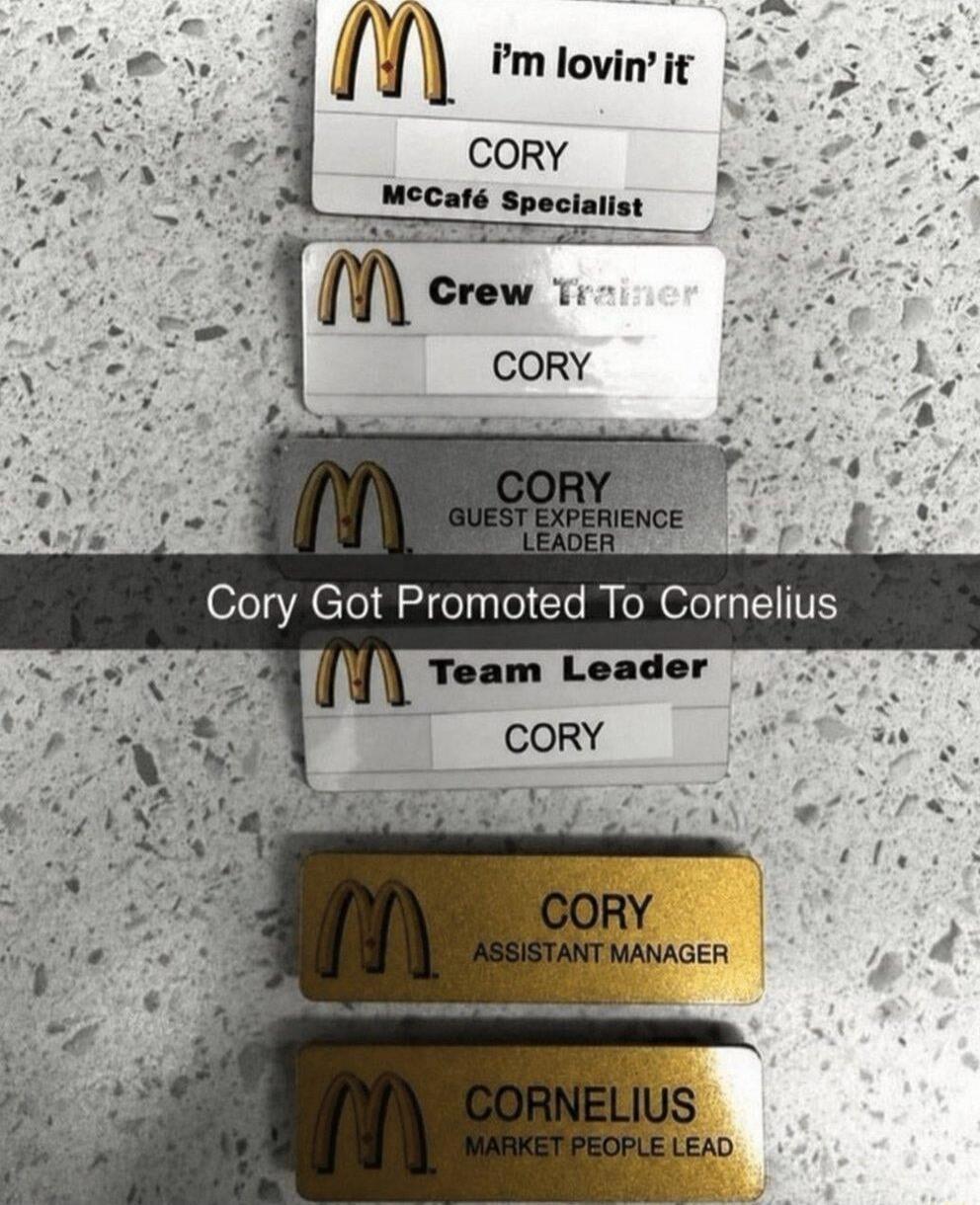 The image displays a series of McDonald's name tags, showing an employee named Cory progressing through various roles: McCafé Specialist, Crew Trainer, Guest Experience Leader, Team Leader, and Assistant Manager. The final name tag, for the role of Market People Lead, shows the name 'Cornelius' instead of 'Cory', with a caption stating 'Cory Got Pr