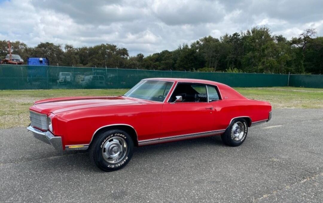 A red classic car, possibly a Chevrolet Monte Carlo, is parked on an asphalt surface. The tires have 'COOPER COBRA RADIAL G/T' written on them. In the background, there is a green fence and trees under a cloudy sky.