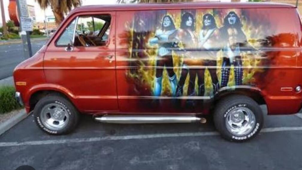 A vintage red van with custom airbrushing of the rock band KISS members in their stage costumes and makeup, surrounded by flames. The van has chrome wheels and side steps, parked on asphalt.