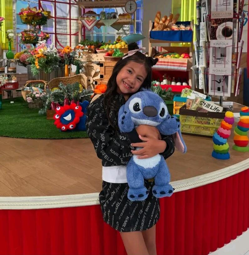 A young girl smiles while hugging a blue Stitch plush toy. She is wearing a black outfit with 'KARL LAGERFELD' text. The background features a colorful set with flowers, fruits, bread, and magazines titled 'FOOD GATHER', 'KINFLK', 'NORDIC', and 'Still Life'.