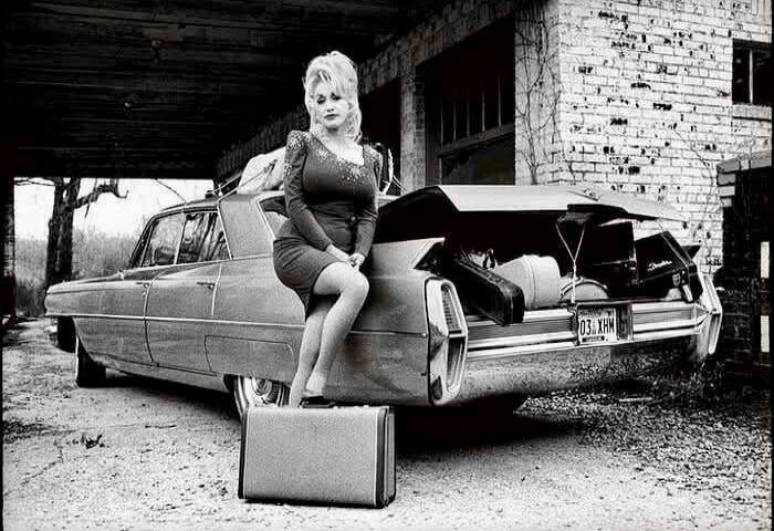 Dolly Parton sitting on the trunk of a classic car with an open trunk, a suitcase on the ground, and a brick building in the background. The license plate reads '03 XHM'.