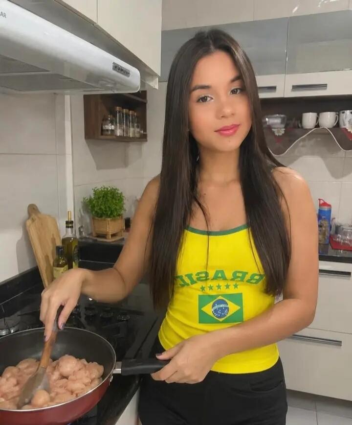 A young woman in a kitchen is stirring raw chicken pieces in a frying pan on a stove. She is wearing a yellow tank top with the Brazilian flag.