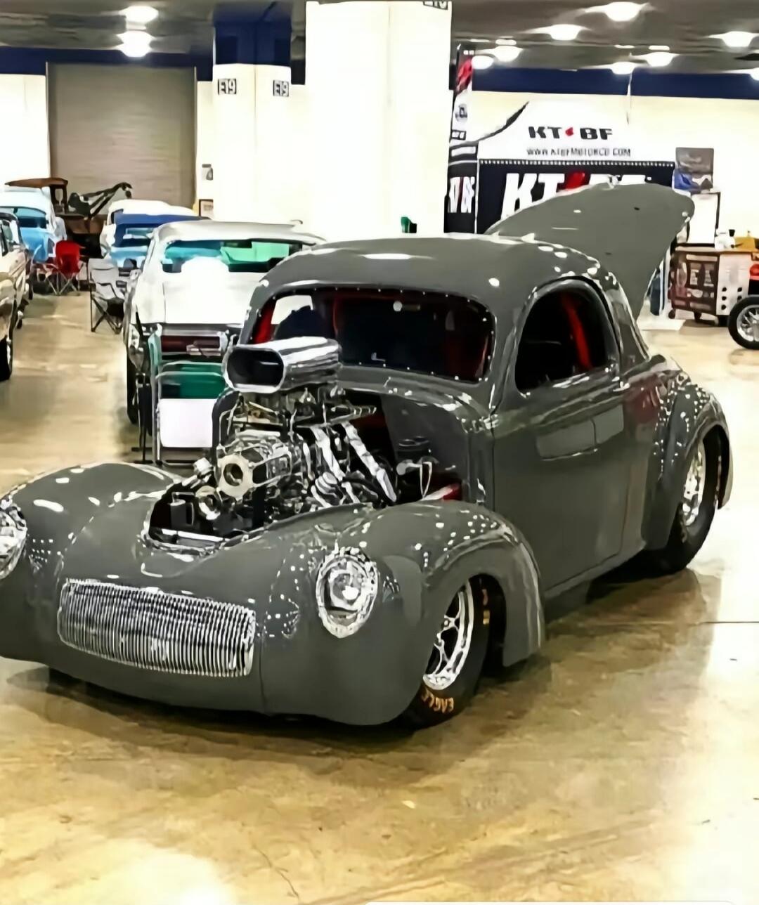 Grey classic hot rod with exposed engine at an indoor car show.