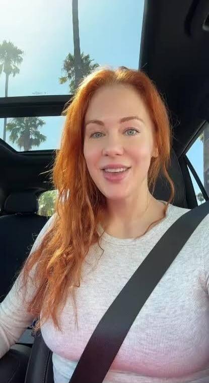 A red-haired woman with blue eyes is smiling in a car, wearing a seatbelt and a light-colored long-sleeve shirt. Palm trees are visible in the background through the window.