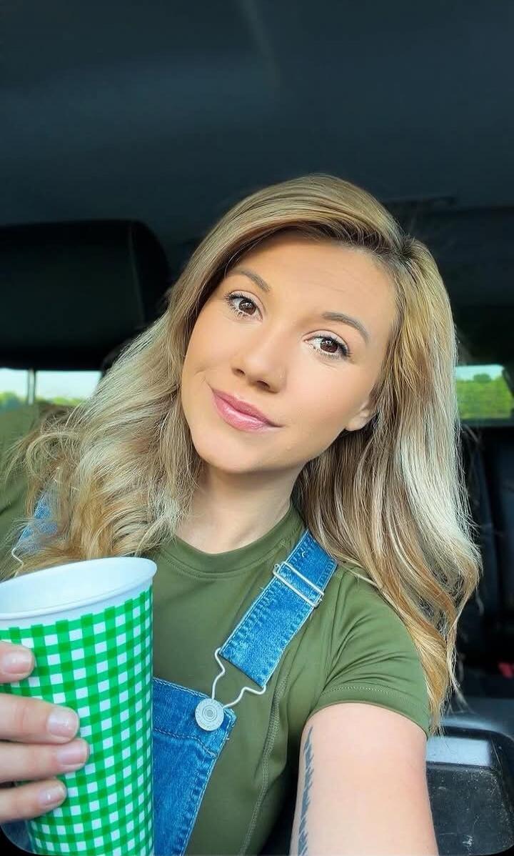 A blonde woman in a green t-shirt and denim overalls is smiling and holding a green and white checkered cup. She appears to be inside a car.