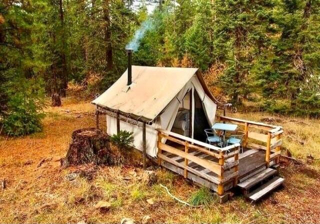 A cozy canvas tent with a chimney emitting smoke, set on a wooden deck with a table and chairs, surrounded by a dense forest with tall evergreen trees and dry ground foliage. A tree stump is visible nearby.