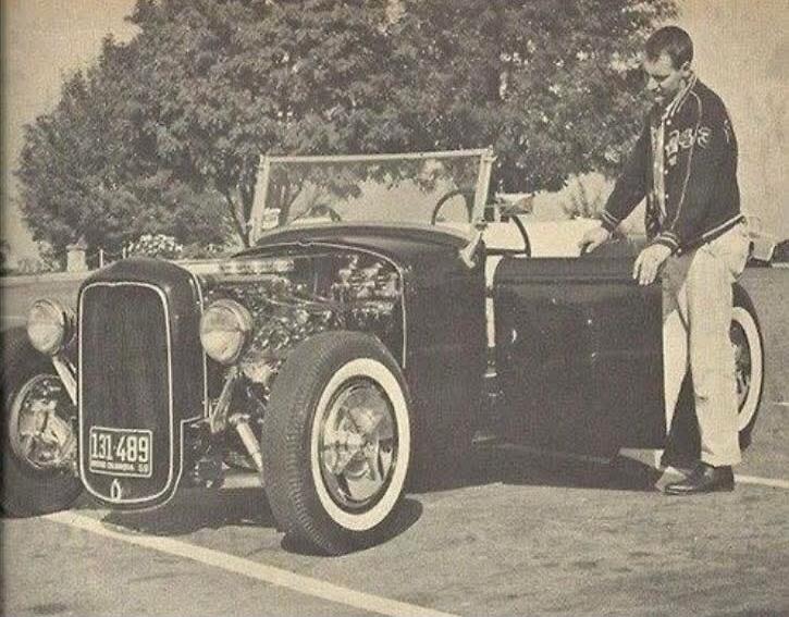 A black and white photo of a man standing next to a vintage hot rod car. The car has whitewall tires and an exposed engine. The license plate reads '131 489 SO. DAKOTA 55'.