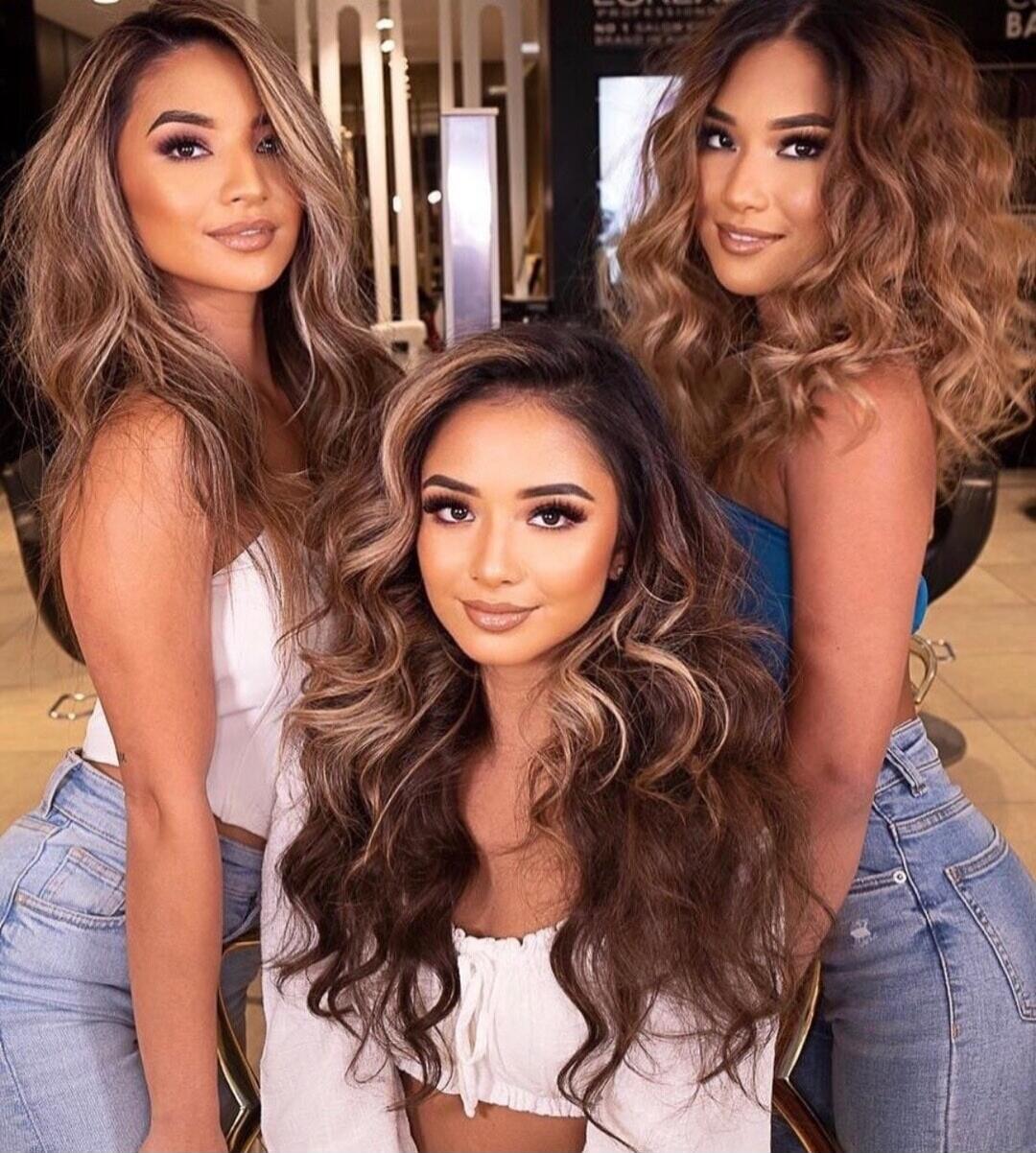 Three women with long, wavy, styled hair, wearing makeup and fashionable tops, posing in what appears to be a salon setting.
