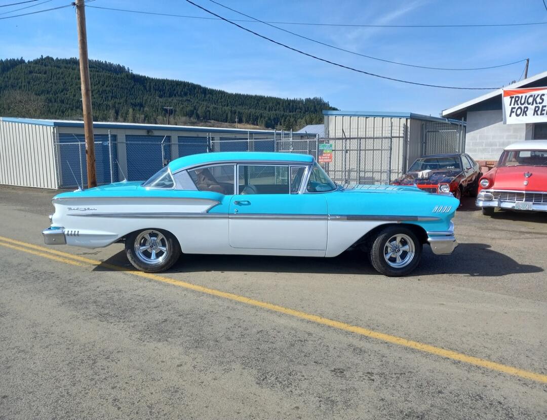 A vintage two-tone light blue and white car, possibly a Chevrolet Bel Air, is parked on an asphalt road. In the background, there are storage units, a utility pole, and a forested hill. Other classic cars are visible. A sign partially reads 'TRUCKS FOR RENT'.