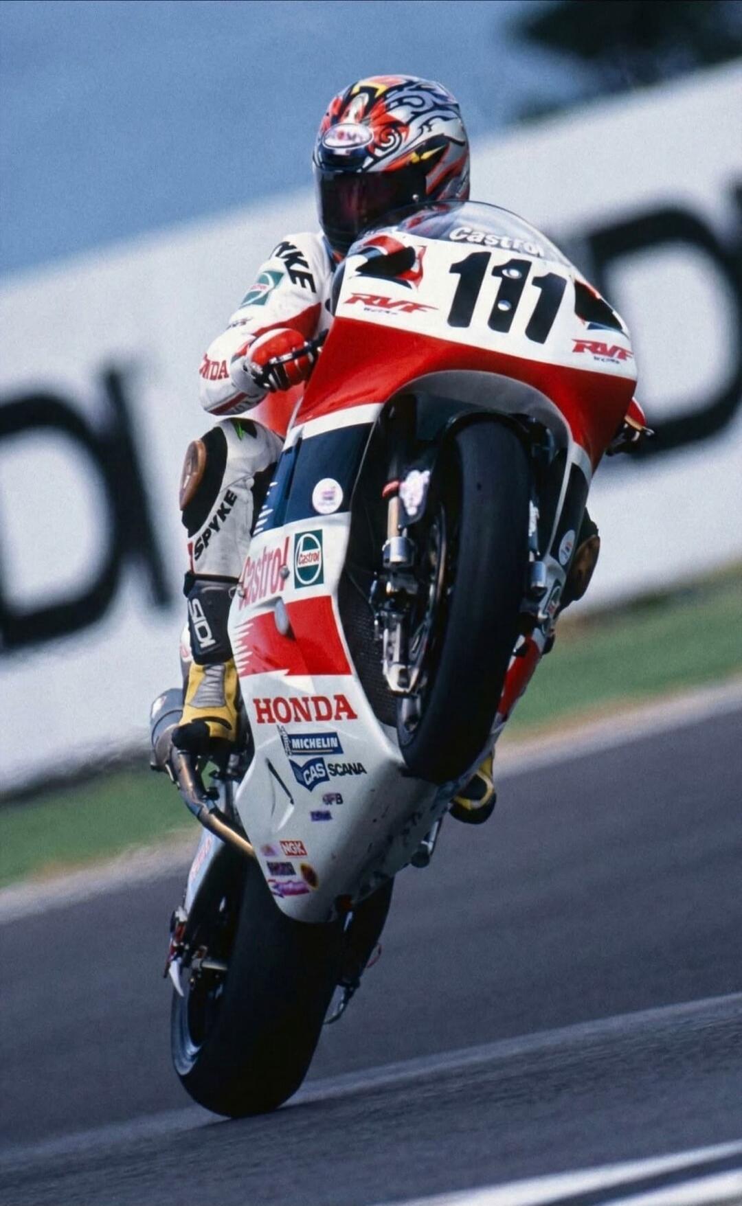 A professional motorcyclist on a Honda RVF bike, number 111, performing a wheelie on a race track. The rider is wearing a racing suit with 'SPYKE' and 'Castrol' logos, and a helmet with 'BELL' visible. The bike features 'HONDA', 'MICHELIN', 'CAS SCANA', and 'NGK' sponsor decals.