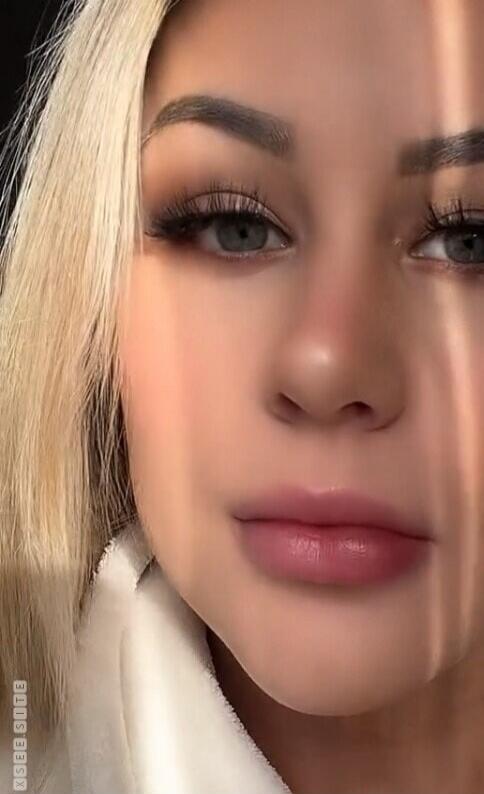 Close-up of a woman's face with blonde hair and makeup, wearing a white robe. Sunlight creates vertical stripes on her face. Watermark 'XSEE SITE' is visible.
