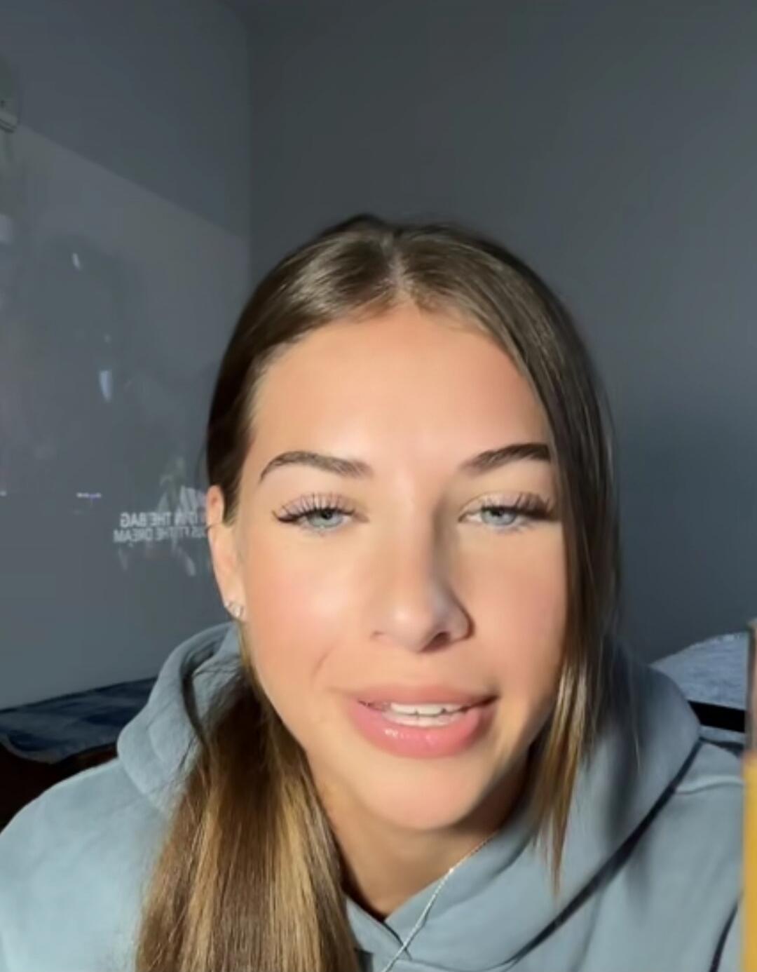 A young woman with long brown hair wearing a gray hoodie speaks to the camera in a plain room.
