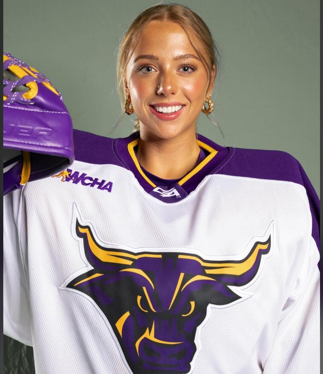 A young woman smiling, wearing a white, purple, and yellow hockey jersey with a bull logo and 'WCHA' text. She is holding a purple hockey glove with 'JER' visible.