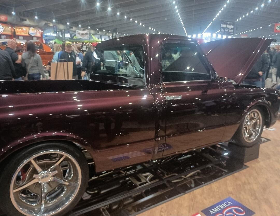 The image shows a custom-built pickup truck with a deep purple metallic paint job. The hood is open, revealing the engine bay. The truck has chrome rims and a lowered suspension. It appears to be on display at a car show or exhibition, with people in the background. There is a sign on the floor that says 
