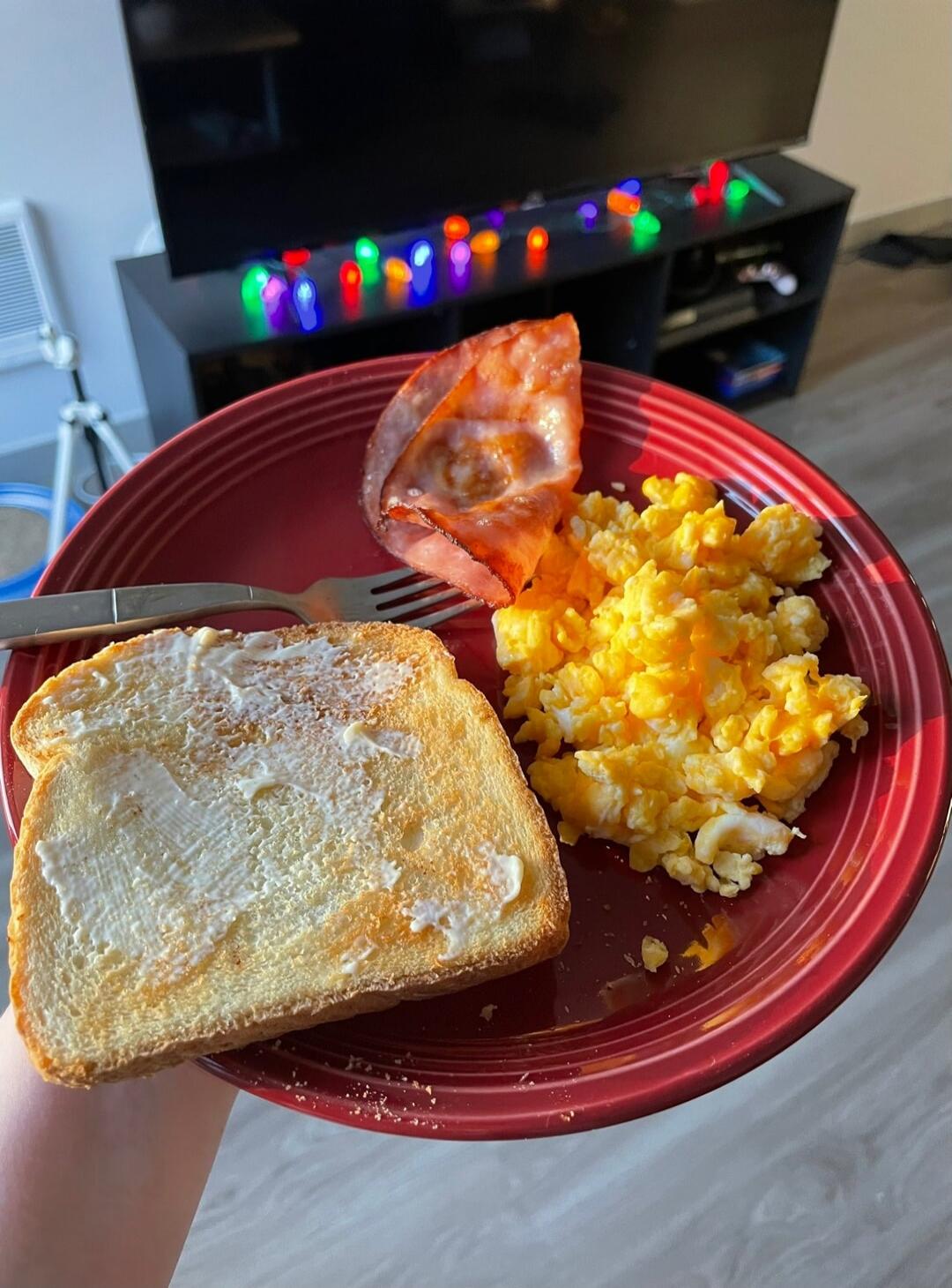 Buttered toast on a red plate with scrambled eggs and a slice of tomato. In the background, a TV stand with colorful string lights.