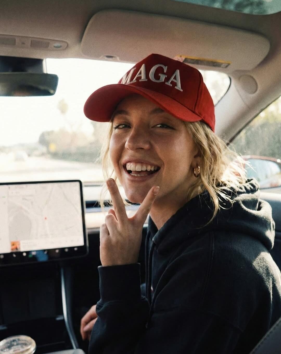 A young woman in a car, smiling and making a peace sign. She is wearing a red baseball cap with 'MAGA' written on it and a black hoodie. A car's navigation screen is visible in the background.