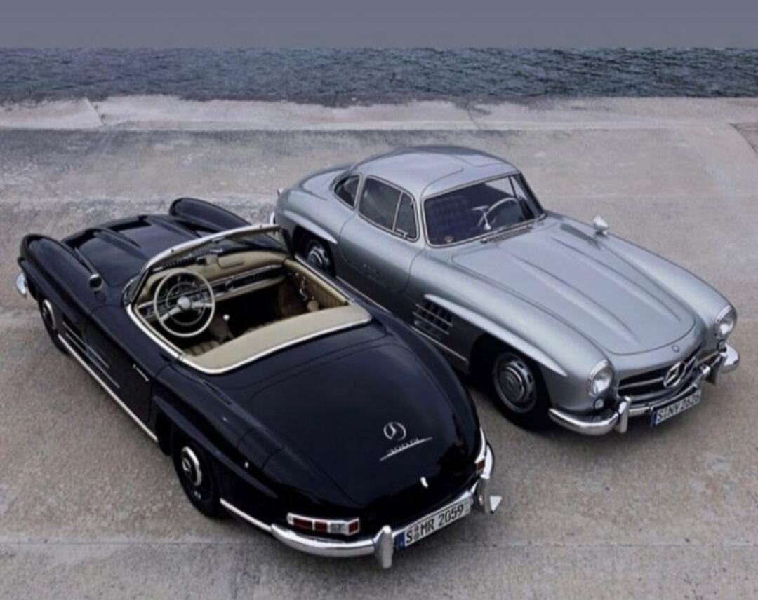 Two classic Mercedes-Benz 300 SL cars, a black convertible and a silver coupe, parked by the sea.