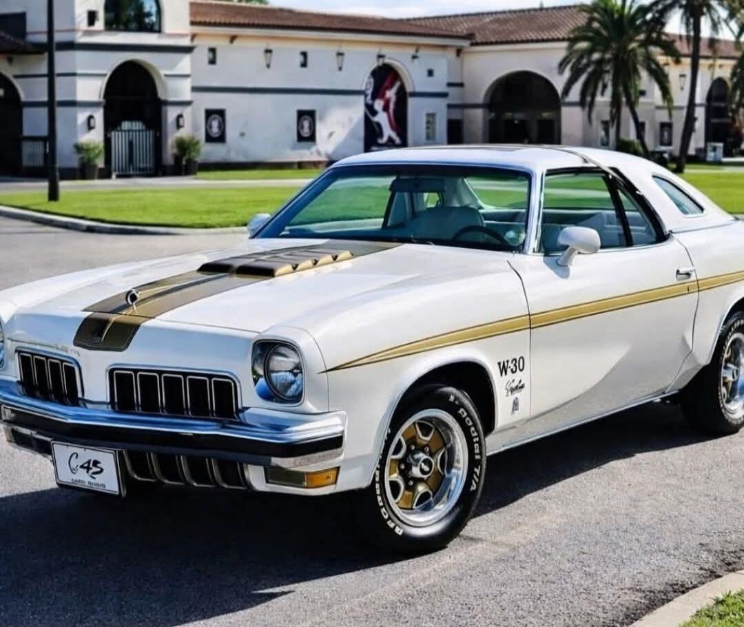 A classic white Oldsmobile Cutlass W-30 with gold racing stripes and a hood scoop is parked on a paved road. The car features BFGoodrich Radial T/A tires and a license plate with 'C45' and 'Little River'. In the background, there's a large building with arches and palm trees.