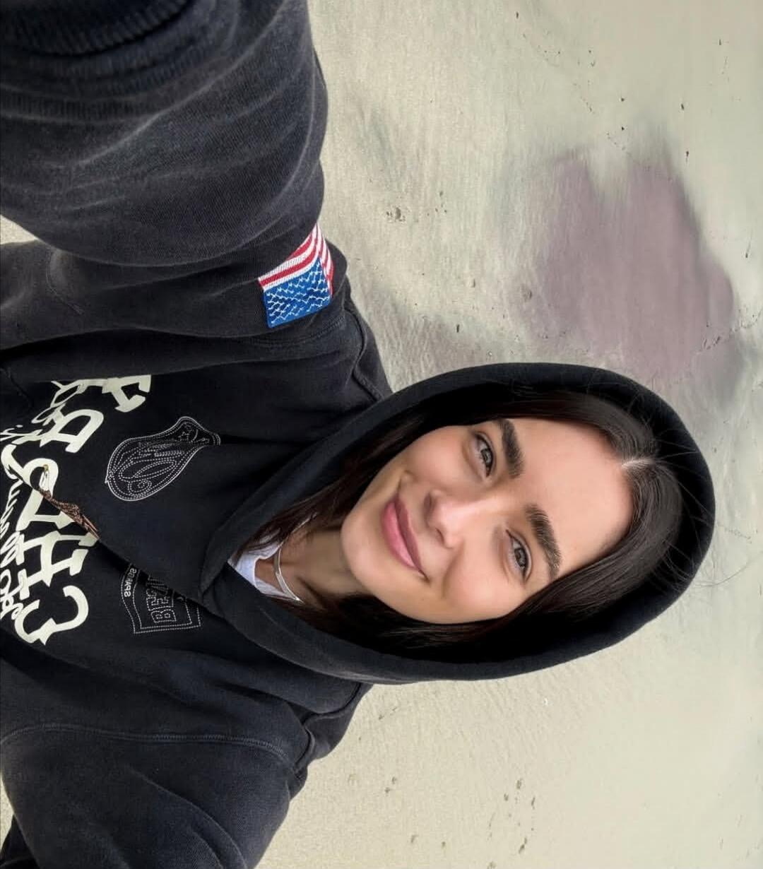 A woman in a black hoodie with white graphic text on it. The text is partially visible but unreadable.