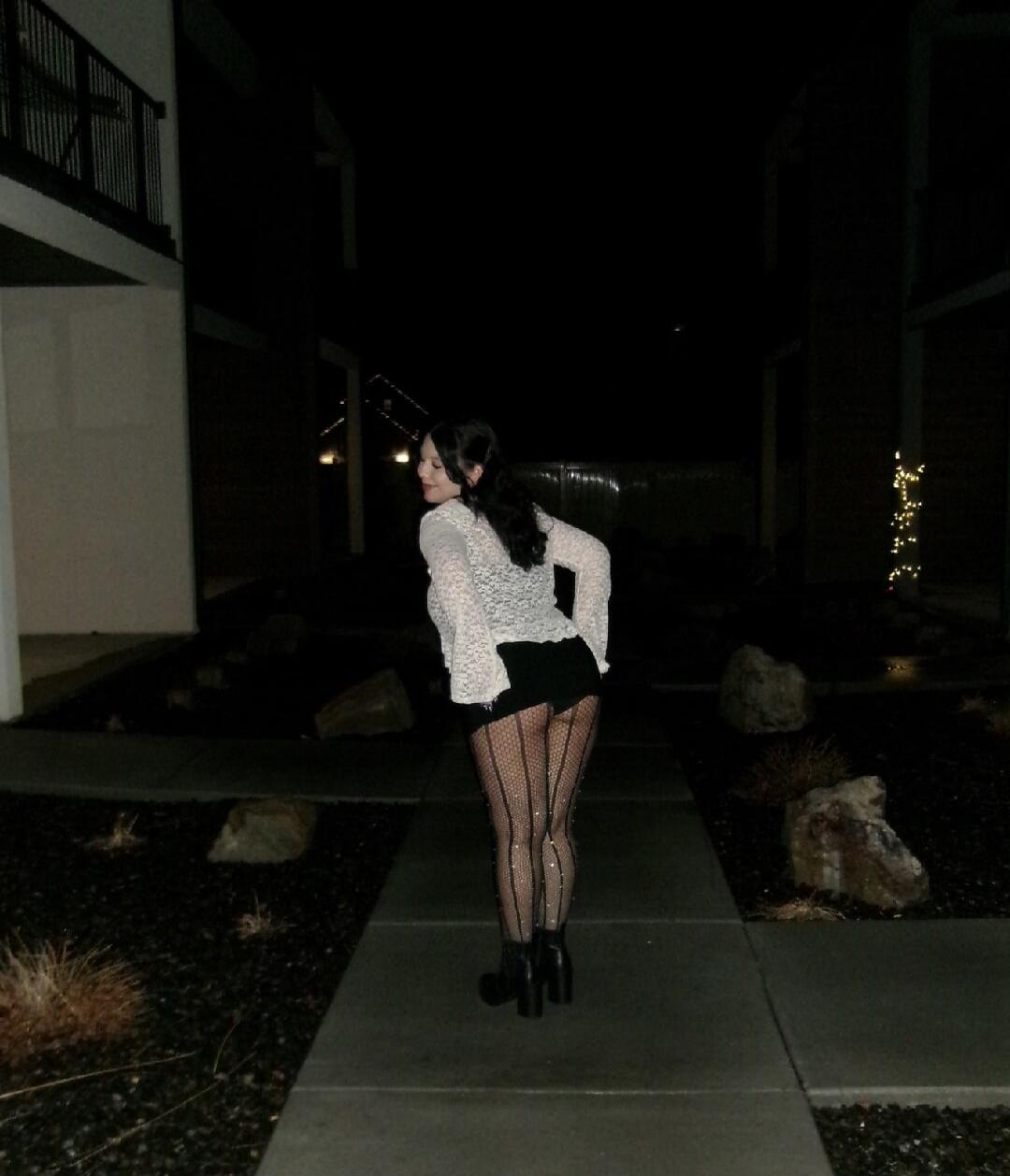 Woman posing outdoors at night wearing a white lace top, black short bottom, fishnet tights, and high-heeled boots.