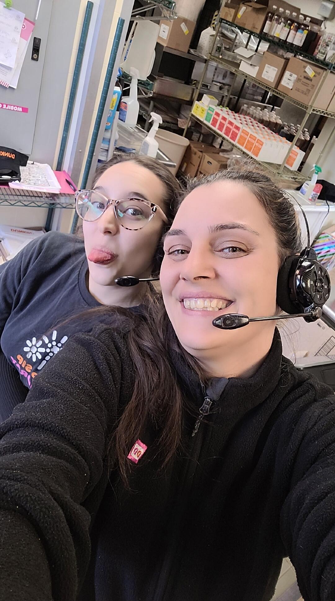 Two women wearing headsets, one smiling and the other making a funny face with her tongue out. They are in what appears to be a backroom or storage area with shelves of products. A 'DUNKIN'' logo is visible on a cap.