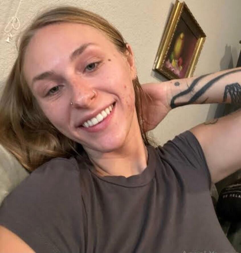 A smiling woman lying down, wearing a gray t-shirt. She has a nose piercing and a tattoo on her arm.