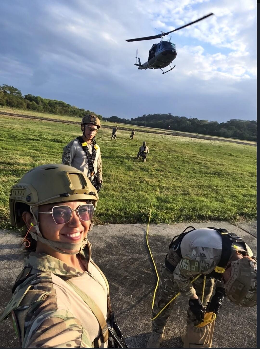 A group of people in military gear are in an open grassy field with a helicopter flying in the background. One woman in the foreground is smiling and taking a selfie, wearing a helmet and sunglasses. Others are seen in the background, some appearing to be preparing for an activity with ropes.