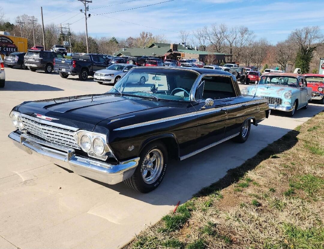 A black vintage car, possibly a 1962 Chevrolet Impala, is parked outdoors on a sunny day. In the background, there are other cars and a building with text 'FRIED CHICKEN' and 'T CREEK'.