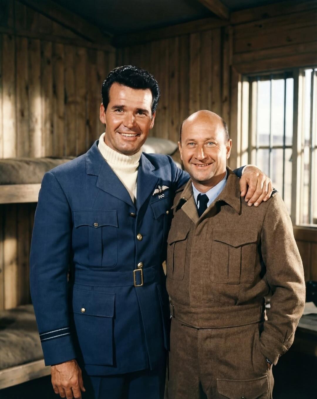 Two men in military-style uniforms, one in a blue uniform with a white turtleneck and the other in a brown uniform, are smiling and posing. They are standing in what appears to be a wooden barracks room with bunk beds visible in the background.