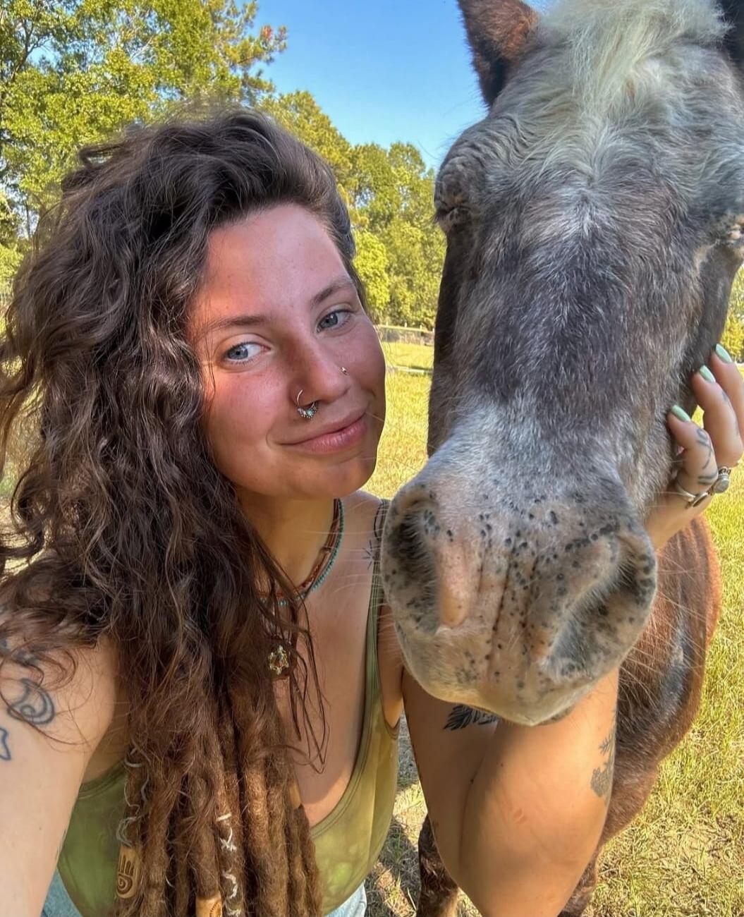 A woman with long curly hair and dreadlocks takes a selfie with a horse in a sunny outdoor setting.