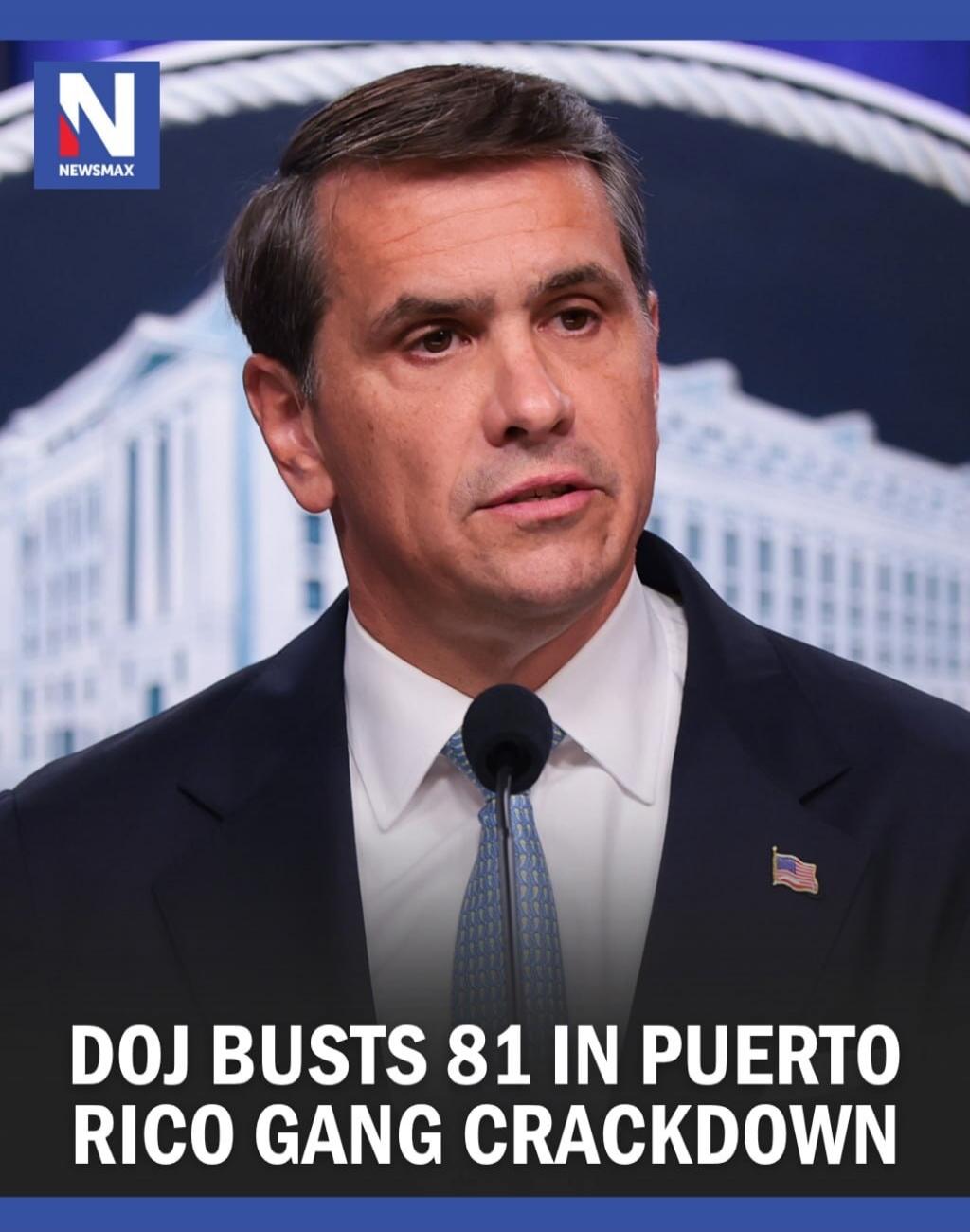 N NEWSMAX DOJ BUSTS 81 IN PUERTO RICO GANG CRACKDOWN. A man is speaking at a podium, likely delivering a news report or statement regarding the Department of Justice's actions.